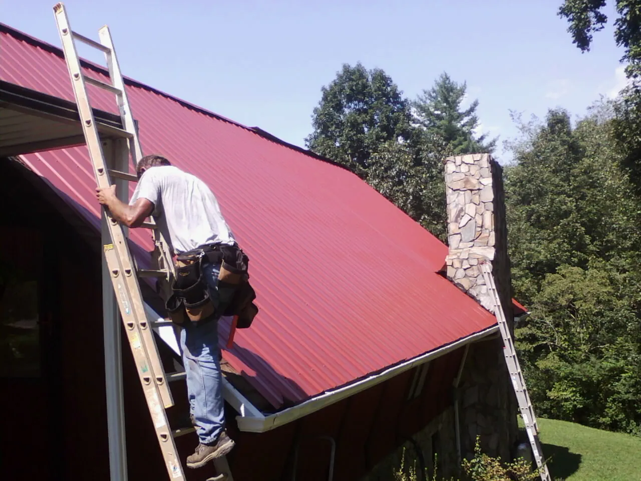 Roofer on a ladder beside a red metal roof and stone chimney — Soffit & Fascia Repair by Roofing Brothers in Moulton